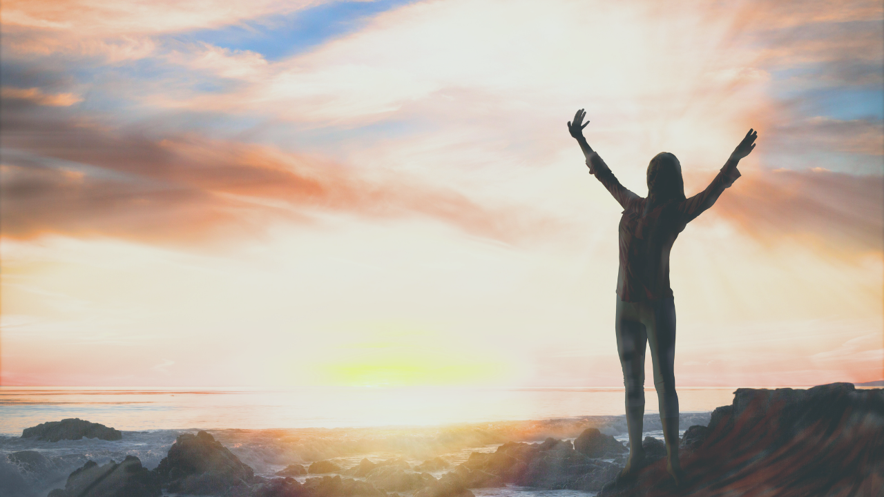 silhouette of a woman standing with her arms raised on a mountain top overlooking a sunset